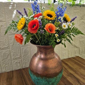Peut inclure: Un vase en cuivre avec une base à patine vert-de-gris contient un arrangement floral vibrant composé de tournesols jaunes, de coquelicots rouges et orange, de delphiniums bleus, de lavande, de marguerites et de fougères.