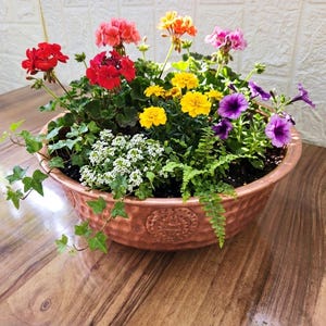 Könnte beinhalten: Eine runde, gehämmert kupferfarbene Pflanzschale, gefüllt mit einer Vielzahl bunter Blumen, darunter rote, rosa, orange, gelbe und violette Blüten, akzentuiert mit grünem Efeu und Farnlaub, was ein lebendiges und dichtes Blumenarrangement ergibt.