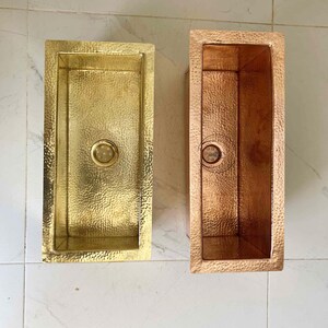 May include: Two rectangular hammered metal sinks. One is gold-toned, the other is copper-toned. Both have a drain in the center. The sinks are displayed on a white tile surface. The sinks are likely for a kitchen or bathroom.