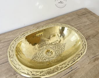 May include: A gold oval-shaped bathroom sink with an intricate floral design etched into the surface. The sink is sitting on a wooden countertop.
