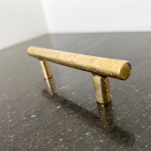 May include: A gold-toned metal cabinet pull with an ornate design. The pull is shaped like a bar with rounded ends.