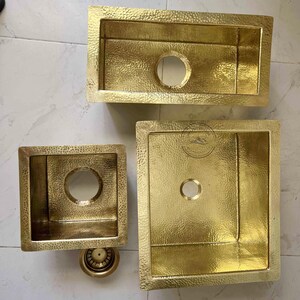 Peut inclure: Trois éviers de cuisine en laiton martelé de différentes tailles. Les éviers sont de forme rectangulaire ou carrée avec un trou d'évacuation circulaire. Le laiton a une surface texturée et une couleur dorée chaude. Une crépine de vidange en laiton est également visible.