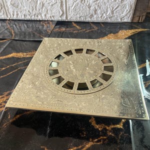 May include: A square drain cover in a gold tone, featuring an elaborate design and a circular grate in the centre.