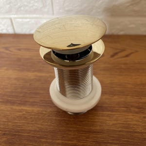 Peut inclure: Un bouchon de vidange pop-up en or pour un évier. Le bouchon a un dessus rond et plat et une base cylindrique filetée.