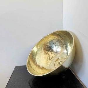 Peut inclure: Un évier en laiton martelé de forme ovale et arrondie. L'intérieur présente une surface texturée et un drain. L'évier est positionné contre un mur blanc et un comptoir noir.