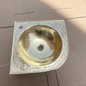 Puede incluir: Un lavabo de esquina de metal dorado martillado con un borde grabado y ornamentado. El lavabo tiene una cubeta circular y un orificio de desagüe. El borde presenta un diseño floral y de volutas detallado.