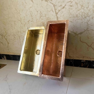 May include: Two rectangular hammered metal sinks. One is gold-toned, the other is copper-toned. Both have a drain in the centre and a textured surface. The sinks are angled, showcasing their interior and exterior details.