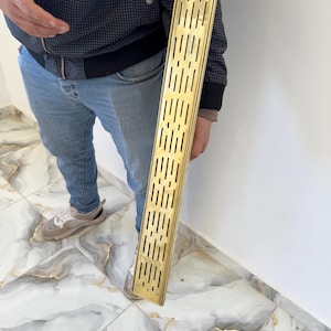 Puede incluir: Una rejilla de drenaje de metal dorada, larga y rectangular, con una serie de pequeños agujeros espaciados uniformemente.
