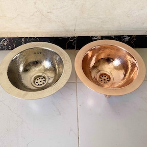 Op de afbeelding: Twee gehamerde metalen wastafels. Eén is zilverkleurig, de andere koperkleurig. Beide hebben een ovale vorm met een getextureerd interieur en een ronde afvoer. De wastafels staan op een witte tegelvloer.