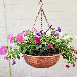Peut inclure: Un bol de suspension en cuivre martelé suspendu par trois chaînes en cuivre assorties et un crochet. Le bol est rempli de fleurs de pétunias vibrantes dans des tons de rose, violet et blanc, entremêlées de feuillage vert et de terreau sombre.