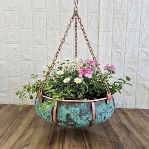 Puede incluir: Una maceta colgante redonda con un acabado de pátina en color verde azulado y cobre, suspendida por tres cadenas de metal en tono cobre. El cuenco está lleno de pequeñas margaritas blancas y flores rosas con follaje verde en los bordes.