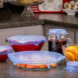 May include: A collection of red and clear glass bowls and a mason jar, each covered with blue silicone lids. The bowls contain food items like oranges and peaches. The jar is filled with a dark liquid.