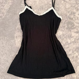 May include: A black camisole featuring a V-neckline trimmed with white lace and thin spaghetti straps. The garment has a simple, sleeveless design with a straight hemline, providing a clean and straightforward appearance for this basic piece of clothing.