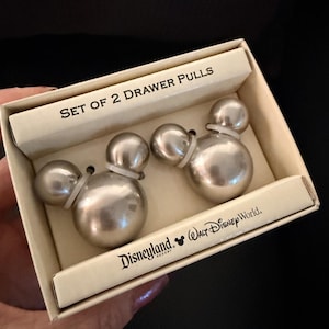 May include: A set of two silver drawer pulls in a box. The drawer pulls are shaped like Mickey Mouse heads. The box is cream-colored and has the text "SET OF 2 DRAWER PULLS" and the Disneyland Resort and Walt Disney World logos.