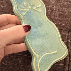 Op de afbeelding: Een lichtblauwe keramische katvormige schaal. De schaal heeft een verhoogde omtrek en details in een lichtere crèmekleur. Het gezicht van de kat heeft twee ronde ogen en een kleine neus. De schaal wordt vastgehouden door een hand met donkerrode nagellak.
