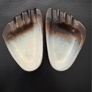 May include: A pair of ceramic ashtrays shaped like footprints. The ashtrays are glazed in a brown and white color scheme.
