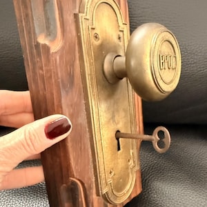 Puede incluir: Antiguo juego de pomo y placa de puerta de latón montado sobre una tabla de madera. El pomo tiene las letras "BPOD" y una llave está insertada en la cerradura. La madera es de color marrón oscuro.