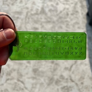 May include: A green plastic keychain with a raised alphabet and symbols. The keychain features the letters A through Z, numbers 1 through 9, and symbols including an arrow pointing up, an arrow pointing down, a square, and a circle.