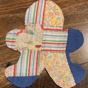 May include: A quilted fabric coaster shaped like a flower with five rounded petals. The surface features a patchwork design of various fabrics, including white with red, green, and blue stripes, a floral print with yellow and orange flowers, and solid blue.
