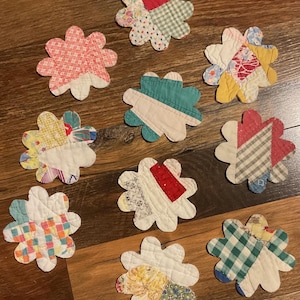 May include: A collection of nine flower-shaped fabric coasters, each featuring a unique patchwork design made from various quilted cotton scraps in assorted colors, patterns, and textures, including gingham, floral prints, and solid red, teal, and yellow.