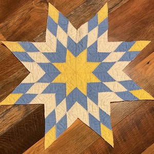 May include: A handmade star-shaped quilt with a yellow center and alternating blue, white, and yellow diamond patterns. The quilt is made of fabric and has a quilted texture. The star is displayed on a wooden surface.
