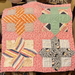 May include: A square patchwork quilt featuring a pink floral background fabric. The quilt is composed of four large star-shaped blocks in orange, light green, multi-colored stripes, and a black and white zebra print with small fruit motifs.