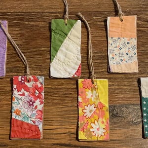 May include: A collection of handmade fabric bookmarks with various patterns and colors. Each bookmark is rectangular, featuring patchwork designs with floral, geometric, and abstract motifs. They have a rustic, homespun aesthetic, with twine attached for hanging.