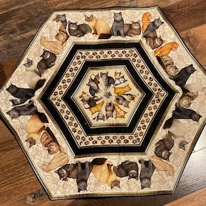 May include: A hexagonal fabric mat featuring a repeating pattern of various cat breeds in shades of brown, gray, orange, and black. The center displays a smaller hexagon with cats arranged in a circle, surrounded by a border of paw prints and floral motifs.