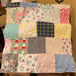 May include: A patchwork quilt with a variety of fabric patterns and colors. The quilt features squares of floral, plaid, and solid fabrics in shades of pink, blue, green, yellow, and gray. The quilt is likely handmade and has a vintage aesthetic.