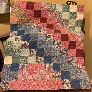 May include: A patchwork quilt with a variety of patterned squares in shades of red, blue, green, and cream. The quilt is made of fabric squares sewn together in a grid pattern. The quilt is draped over a wooden surface.