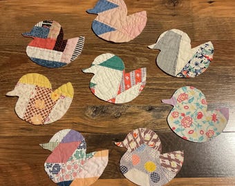 May include: Seven duck-shaped fabric ornaments crafted from various quilted textile scraps. Each piece features a unique patchwork design with diverse patterns including floral, geometric, and solid colours in shades of blue, pink, yellow, and white.