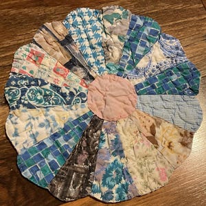 May include: A handmade, circular quilted item with a pink center and petal-like sections in various patterned fabrics. The fabrics include floral, geometric, and paisley designs in shades of blue, green, beige, and brown. The item is likely a trivet or decorative piece.