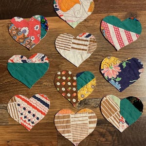 May include: Nine handmade fabric heart ornaments in various patterns and colors, including floral, striped, and patchwork designs. The hearts are arranged on a wooden surface, showcasing a rustic, vintage aesthetic.