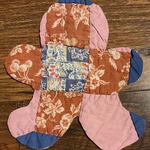 May include: A quilted fabric piece with a flower-like shape, constructed from patchwork squares. The design features a central blue and white patterned square surrounded by rust-colored floral fabric, solid pink sections, and rounded blue edges.