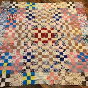 May include: A handmade quilt with a patchwork design. The quilt features a grid pattern of squares in various colors and patterns, including floral, plaid, and solid fabrics. The color palette includes red, blue, pink, and brown.