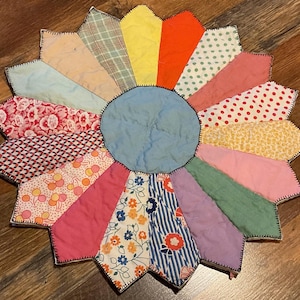May include: A colorful, handmade quilted trivet in a flower shape. The trivet features a light blue center and petal-shaped sections in various patterns and colors, including floral, polka dots, and stripes. The edges are finished with black stitching.
