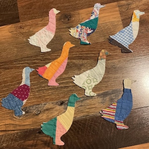 May include: Seven fabric goose-shaped appliques featuring various patchwork patterns and colors, including blue gingham, floral prints, stripes, and solid pink, orange, yellow, and green sections, all stitched with visible quilting lines throughout.