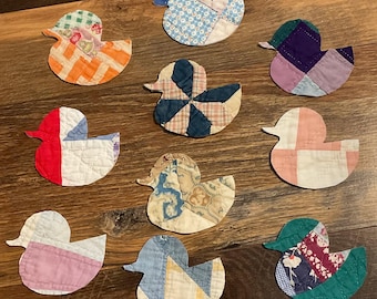 May include: A collection of nine fabric duck-shaped appliqués, each crafted from various quilted textile scraps in diverse colours and patterns, including orange, blue, red, pink, purple, and floral prints, arranged in a scattered formation.