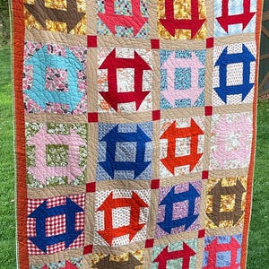 Op de afbeelding: Een handgemaakte quilt met een herhalend patroon van kleurrijke blokken. Elk blok heeft een centraal vierkant met pijlvormige vormen in verschillende kleuren, waaronder rood, blauw en bruin, tegen een beige achtergrond. De quilt is afgezet met een roestkleurige stof.
