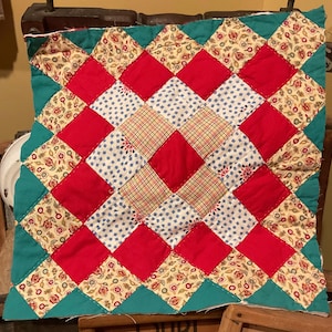 May include: A handmade quilt with a diamond pattern. The quilt features red, white, and floral patterned fabric squares. The border is teal. The quilt is likely made of cotton and is a vintage item.