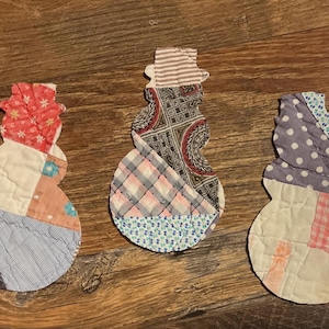 May include: Three snowman-shaped fabric ornaments crafted from various quilted patches. Each figure features a distinct combination of patterned textiles, including floral, plaid, polka dot, and geometric prints in shades of red, blue, grey, and white.