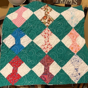 May include: A square quilt featuring a dark green background with white stitched swirl patterns. The quilt is composed of diamond-shaped patches in various patterns, including pink stripes, brown floral, blue dots, red paisley, and multi-colored floral prints.