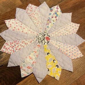 May include: A handmade, quilted flower-shaped trivet. The petals are made of various patterned fabrics in shades of white, pink, blue, and yellow. The center features a circular design with black and white swirls and a red accent.