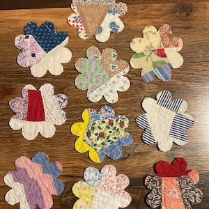 May include: Nine handmade, quilted flower-shaped coasters. Each coaster features a unique patchwork design with various fabric patterns and colors, including blue, red, yellow, and pink. The coasters are arranged on a wooden surface.