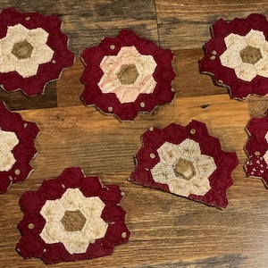 May include: A set of six fabric coasters featuring a hexagonal patchwork design. Each coaster is constructed from small fabric pieces in deep burgundy, cream, and tan, arranged in a floral-like pattern with scalloped edges and visible stitching.