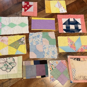 May include: A collection of colorful, handmade quilt blocks in various shapes and patterns. The blocks feature a mix of fabrics in shades of yellow, pink, blue, and green. Some blocks have geometric designs, while others feature floral or butterfly motifs.