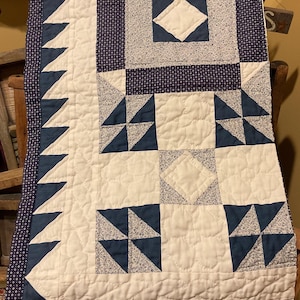 May include: A handmade quilt with a patchwork design. The quilt features a white background with navy blue and light blue patterned squares and triangles. The border is navy blue with a small white dot pattern. The quilt is displayed on a wooden chair.