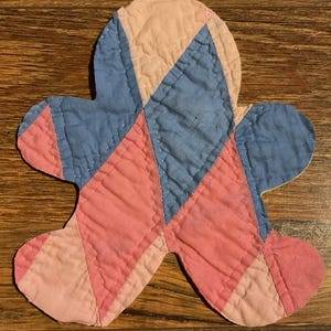 May include: A quilted fabric ornament shaped like a gingerbread person, constructed from triangular patches of light pink, dusty rose, and muted blue fabric. The piece features visible white stitching along the seams of the geometric pattern.