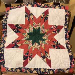 May include: A square quilted textile featuring a central eight-pointed star pattern. The star is composed of dark green, solid red, and floral-patterned fabric in shades of red, yellow, and green. White fabric squares fill the corners.
