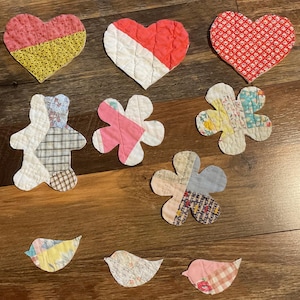May include: A collection of handmade fabric appliqués in various shapes, including hearts, flowers, a teddy bear, and birds. The appliqués feature a patchwork design with a variety of colorful patterned fabrics, including floral and geometric prints.